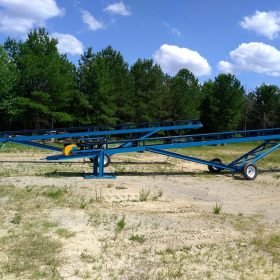 A blue metal conveyor system stands idle on a grassy, sandy surface, surrounded by trees under a clear, partly cloudy sky.