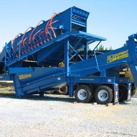 A blue industrial screening machine with multiple vibrational screens on a trailer, surrounded by trees.