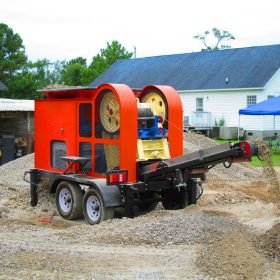 A mobile rock crusher processes gravel, discharging through a conveyor.