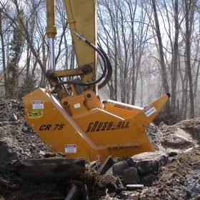 Excavator attachment labeled "CRUSH-ALL CR 75" actively breaks concrete debris amidst a wooded area.