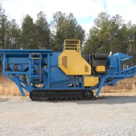A large, industrial mobile crusher rests on gravel ground, featuring blue and yellow metal components.